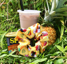 Load image into Gallery viewer, A cotton scrunchie featuring Hawaiian local childhood favorites such as pineapple, poke bowl, spam musubi, milk tea, and shave ice on a yellow background standing in front of a can of spam, a pineapple, and a milk tea sitting in the grass.
