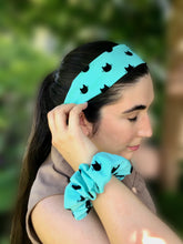 Load image into Gallery viewer, A dark-haired woman wearing a blue-teal cotton flat headband with cute and simple cat face designs, paired with a matching scrunchie on her wrist, perfect for adding a playful touch to any outfit.
