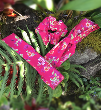 Load image into Gallery viewer, An image of a knotted headband, a flat headband, and a scrunchie all made from the same fabric a floral cotton fabric featuring green leaves and pink and purple flowers on a pretty pink background.
