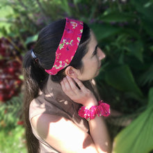 Load image into Gallery viewer, A dark-haired woman wearing a pink flat cotton headband with a lovely floral pattern featuring green leaves and pink and purple flowers on a pretty pink background, adding a touch of colorful charm to her outfit.
