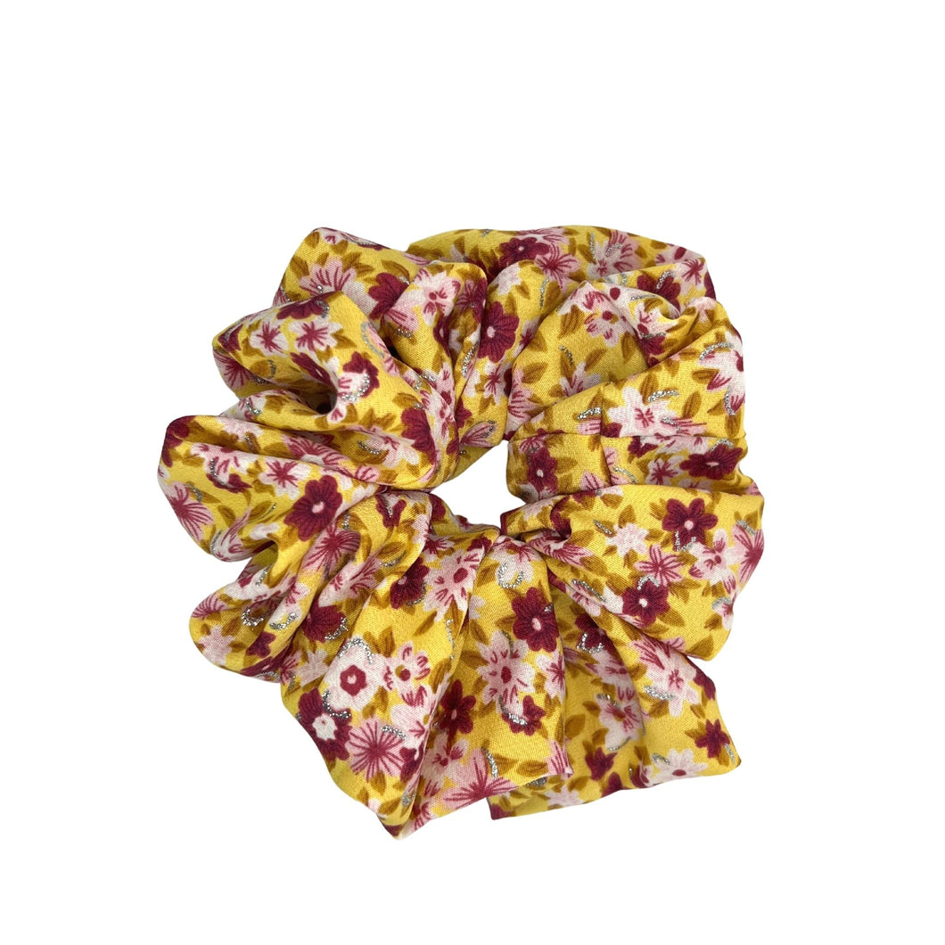A photo of an XXL scrunchie made from a yellow fabric featuring different shades of pink flowers and a touch of silver sparkle, perfect for adding a fun and vibrant pop of color to any hairstyle.