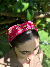Load image into Gallery viewer, A dark-haired woman wearing a pink knotted cotton headband with a lovely floral pattern featuring green leaves and pink and purple flowers on a pretty pink background, adding a touch of colorful charm to her outfit.
