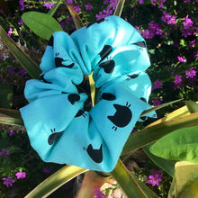 Load image into Gallery viewer, Cotton scrunchie made from blue-teal fabric with cute and simple cat face designs against green plants, perfect for adding a fun and playful touch to any hairstyle.
